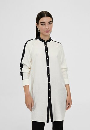 Camisa larga de botones en crema con detalles en negro, que presenta un cuello con solapa, mangas largas y un tejido ligeramente texturizado.