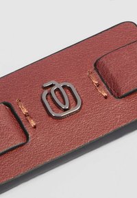 Cinturino in pelle rossa testurizzata con bordi neri, caratterizzato da un logo metallico argentato e cuciture arancioni a contrasto. Due passanti per la chiusura visibili.