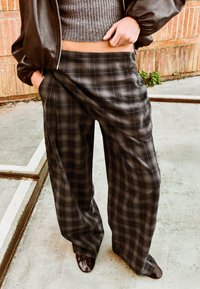 Oseba, oblečena v sive karirane hlače širokih nog, sivi oprijet pulover s kratkimi rokavi, rjav usnjen suknjič in rjavi čevlji, stoji na betonu ob opečnem zidu.