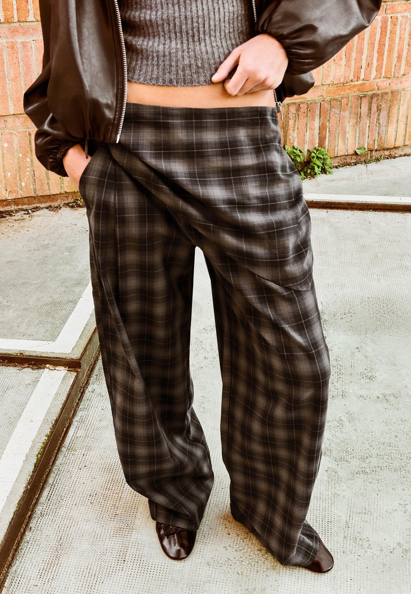 Oseba, oblečena v sive karirane hlače širokih nog, sivi oprijet pulover s kratkimi rokavi, rjav usnjen suknjič in rjavi čevlji, stoji na betonu ob opečnem zidu.