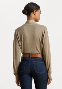 Femme avec les cheveux en chignon portant un haut beige à manches longues rentré dans un jean bleu foncé avec une ceinture marron, vue de dos sur fond blanc.