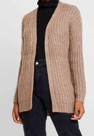 Frau trägt eine beige strukturierte Strickjacke über einem schwarzen Rollkragenpullover und hoch taillierte schwarze Jeans, steht vor einem schlichten Hintergrund.
