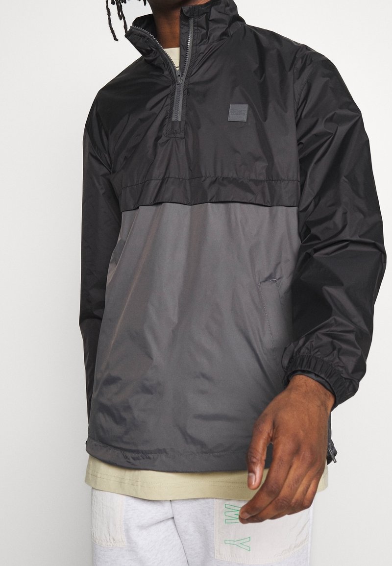 Homme portant un coupe-vent noir et gris à fermeture éclair partielle avec des poignets élastiques, une chemise beige en dessous, et un pantalon clair avec des lettres vertes.