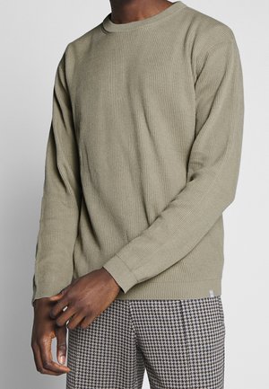 Chemise à manches longues côtelée vert olive avec un col rond et des poignets ajustés, associée à un pantalon à motifs noir et blanc.