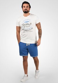 Weißes T-Shirt mit blauer Grafikschrift, kombiniert mit blauen Shorts mit umgeschlagenen Säumen und Kordelzug. Getragen mit weißen Turnschuhen.