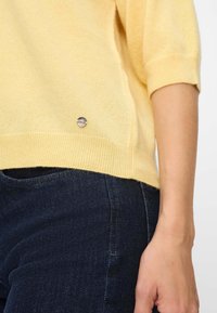 Pull tricot jaune à manches courtes, texture légère, ourlet côtelé et petit détail argenté au-dessus de l'ourlet, assorti à un denim foncé.