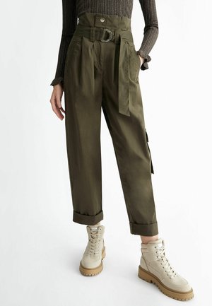 Kvinde iført olivengrønne bukser med høj talje og bælte, med opsmøgede ben og beige snørestøvler, stående med den ene hånd i lommen.