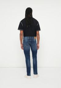 Kék farmernadrág, slim fit szabással, bőrből készült folttal a hátsó részen, öt zsebes kialakítással és kontrasztos varrással. Fekete, rövid ujjú póló felette.