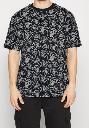 T-shirt noir à manches courtes avec des logos Raiders gris et blancs répétés partout, porté par une personne avec un pantalon noir.