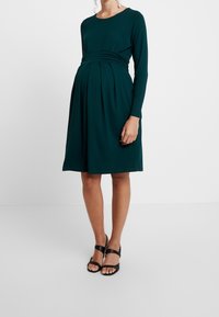 Grön långärmad gravidklänning med rund halsringning, samlad midja och veckad kjol. Tyget har en slät, mjuk textur.