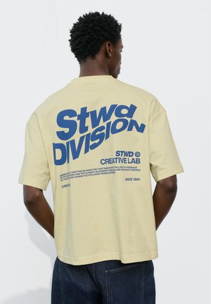Mężczyzna w jasnożółtej, oversize koszulce z dużym niebieskim napisem „Stwd Division” oraz mniejszym logo Creative Lab na plecach, w połączeniu z ciemnymi dżinsami.