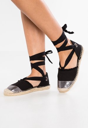 Nogi w czarnych płóciennych espadrylach z srebrnymi czubkami i czarnymi wstążkami zawiązanymi wokół kostek na białym tle podłogi.