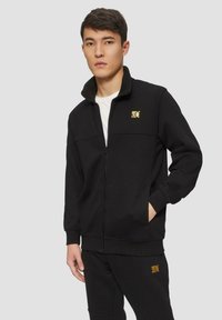 Schwarze Zip-Jacke aus weichem Stoff mit hohem Kragen. Verfügt über ein goldenes Logo auf der Brust und passende schwarze Hosen. Minimalistisches Design.