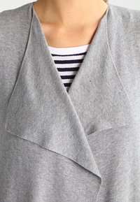 Světle šedý cardigan s volně splývajícím límcem a měkkou pleteninou, přeložený přes bílou a námořnickou pruhovanou košili.