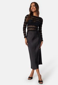 Top in pizzo nero a maniche lunghe con design trasparente e motivi intricati abbinato a un gonna midi nera in satin, dalla texture morbida e silhouette fluida.