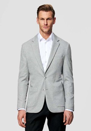 Mężczyzna w jasnoszarej marynarce w kratę, białej koszuli i czarnych spodniach, stojący na tle jednolitego szarego tła.