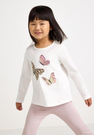 Ragazza giovane con capelli neri lisci che indossa una maglietta bianca a maniche lunghe con tre farfalle colorate e leggins rosa chiaro.