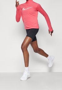 Lyserød langærmet atletisk top, sorte cykelshorts og hvide sneakers. Toppen har et hvidt Nike-logo og sporty designelementer.