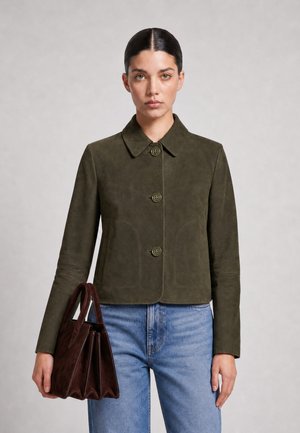Woman with dark hair in a green buttoned jacket and blue jeans holding a dark brown handbag against a plain background.