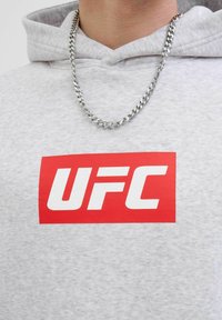 Nahaufnahme einer Person, die einen hellgrauen Hoodie mit einem roten UFC-Logo und einer silbernen Kettenhalskette um den Hals trägt.