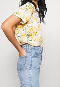 Blouse florale à manches courtes en jaune clair, ornée de fleurs oranges et de feuilles vertes, associée à un jean en denim bleu taille haute.