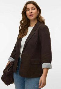 Blazer marron foncé à texture lisse, avec des revers crantés, une fermeture à un bouton et des poches à rabat. En dessous, un haut blanc et un jean bleu clair.