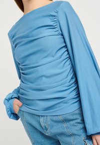 Haut bleu à manches longues avec texture froncée, doté d'un col rond et de poignets élastiqués, associé à un jean en denim clair.