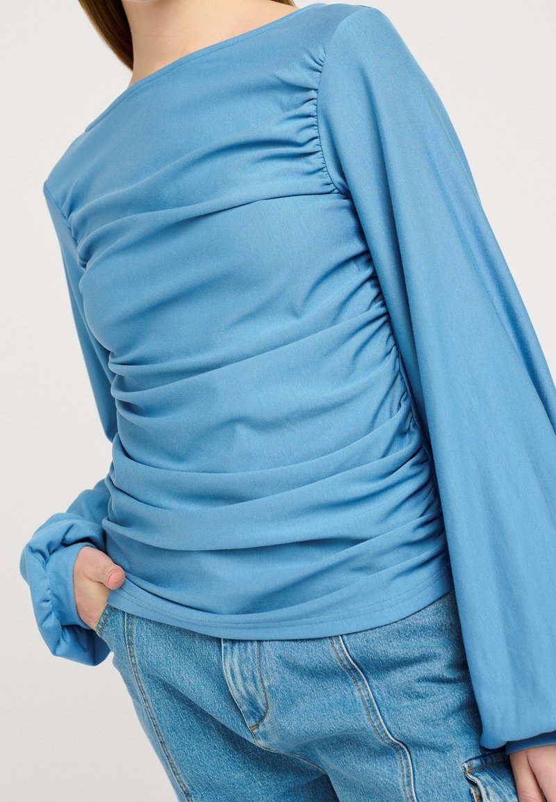 Haut bleu à manches longues avec texture froncée, doté d'un col rond et de poignets élastiqués, associé à un jean en denim clair.