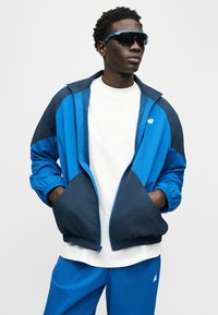 Άνδρας ντυμένος με μπλε και ναυτικό μπουφάν αδιάβροχο, λευκό μπλουζάκι, μπλε παντελόνι και μαύρα αθλητικά γυαλιά ηλίου, με τα χέρια στις τσέπες του μπουφάν, κοιτάει προς τα δεξιά.