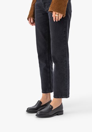 Osoba ubrana w czarne jeansy o kroju cropped, czarne skórzane mokasyny z małym metalowym detalem oraz brązową, teksturowaną bluzkę z długim rękawem.