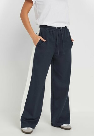 Person iført marineblå vidde bukser med hvide sidelommer, elastisk talje, snøre, hvide sneakers og hvid t-shirt, hænder i lommerne.
