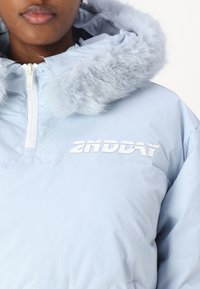 Svetlomodrá prešívaná bunda s kapucňou so syntetickým kožúškom. Má zips a biele logo "2NDDAY" na hrudi. Hladká textúra.