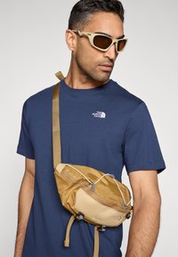 Καφέ τσάντα χιαστί από νάιλον με ρυθμιζόμενο λουράκι, που διαθέτει φερμουάρ και μεταλλικές λεπτομέρειες, φορεμένη πάνω από μπλε σκούρο t-shirt.