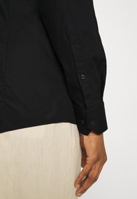 Camisa negra de manga larga con puños abotonados, que presenta una textura suave y un corte entallado, combinada con tela beige clara en la parte inferior.