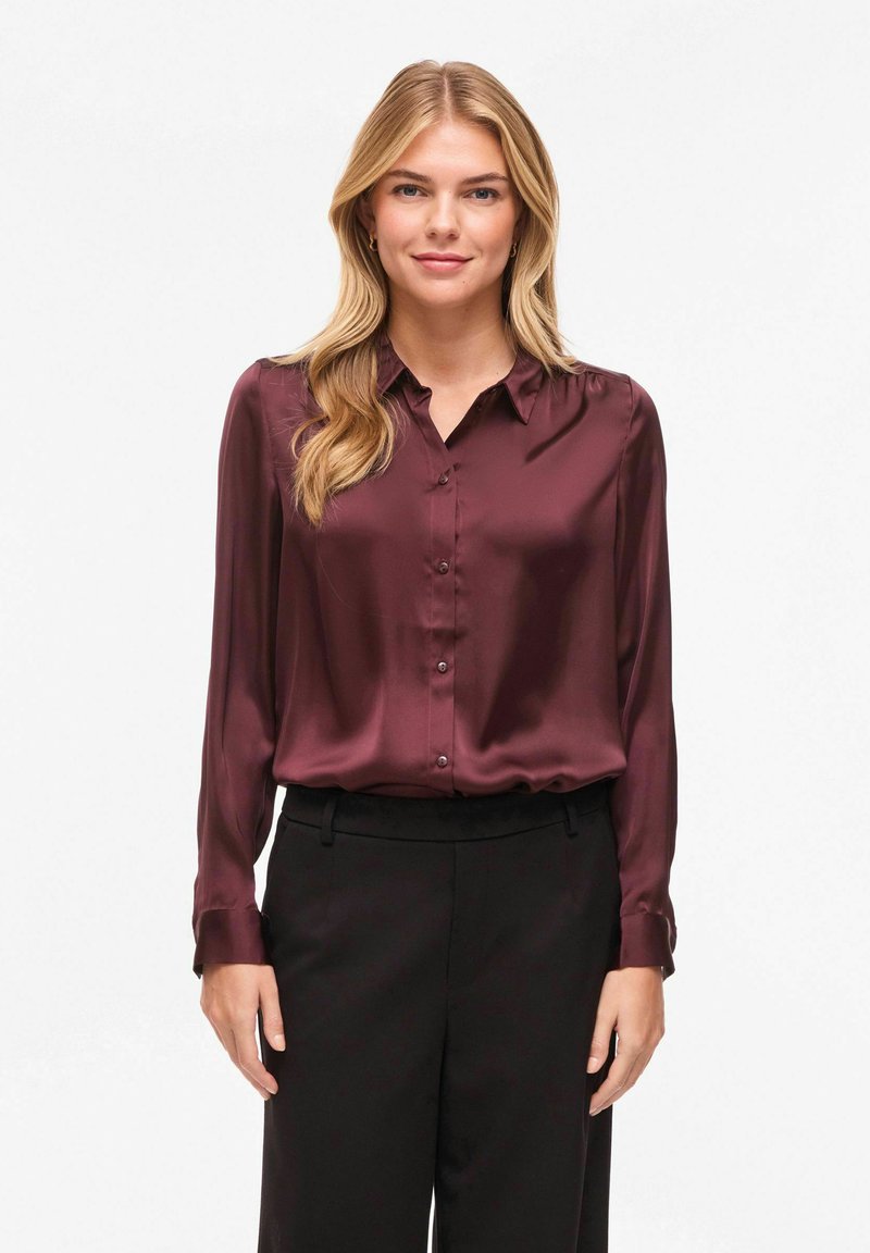 Blouse en satin bordeaux à manches longues, avec patte de boutonnage à l'avant et col. Portée avec un pantalon noir. Texture lisse et finition brillante.