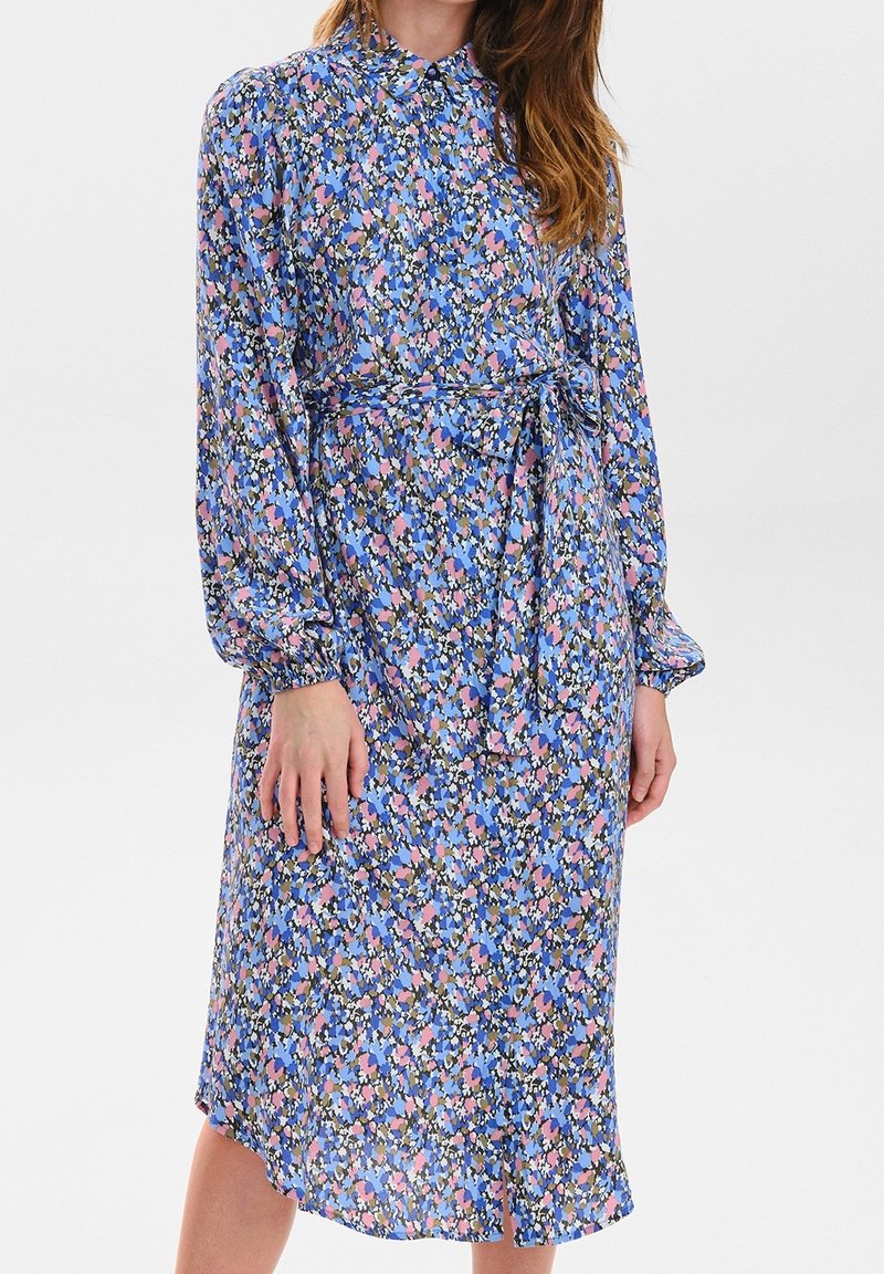 Robe fleurie avec un fond bleu ornée de motifs roses et verts, manches longues, col à boutons et une ceinture assortie.