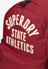 Mochila burdeos con el texto frontal "SUPERDRY STATE ATHLETICS" en grandes letras mayúsculas blancas, con tiradores de cremallera visibles en la parte superior.
