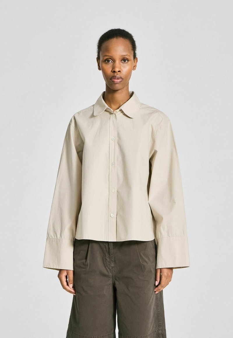 Femme portant une chemise beige oversize à boutons avec larges poignets et un pantalon ample foncé, debout devant un fond clair uni.
