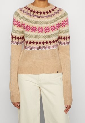 Beige trui met roze, groene en bordeauxrode geometrische en bloemmotieven over de borst en bovenste mouwen, gedragen met crème kleurige broek.