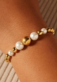 Goud- en wit parelarmband met afwisselend gladde gouden en glanzende parelstenen, afgezet tegen een textuur van huid.