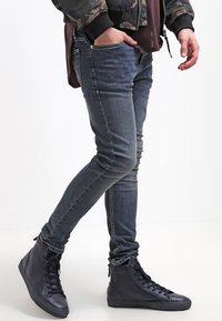 Vaqueros de ajuste ceñido en denim azul oscuro, con cintura de altura media, diseño clásico de cinco bolsillos y costuras visibles. Combinados con zapatillas negras de caña alta.