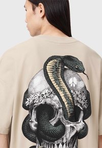 Camiseta beige con un gran gráfico de una serpiente enrollada alrededor de una calavera, con acentos florales y detalles intrincados, textura suave de algodón.