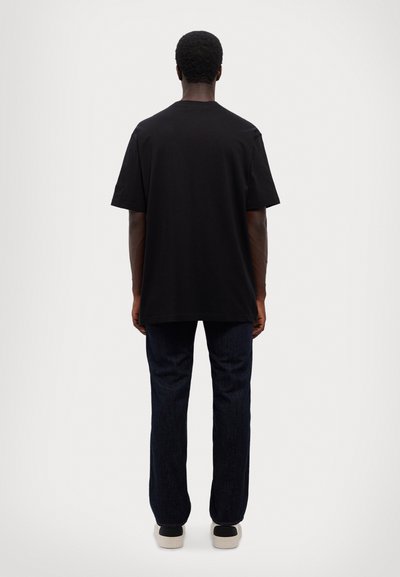 Črna bombažna majica s sproščenim krojem, okroglim izrezom in kratkimi rokavi, v kombinaciji s temno modrimi kavbojkami in črnimi športnimi copati.