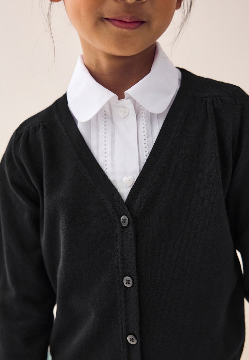 Jeune personne portant une chemise blanche à col avec des boutons en forme de cœur sous un cardigan noir boutonné.