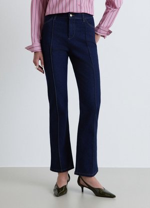 Donna che indossa jeans blu scuro a gamba dritta, camicia rosa a righe a maniche lunghe e scarpe lucide verde scuro con tacco basso.