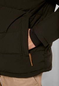 Sort polstret jakke med en synlig sidelomme og detalje med snoretræk. Materialet er glat med en quiltet tekstur og varm udstråling.