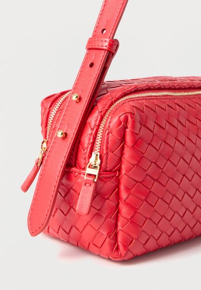 Κόκκινη υφασμάτινη δερμάτινη τσάντα με χρυσή φερμουάρ και ρυθμιζόμενο λουρί σε απλό λευκό φόντο.