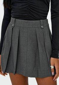 Jupe plissée grise avec une taille haute, ornée de deux boutons. Réalisée en tissu texturé, mettant en valeur des plis structurés et des lignes épurées.