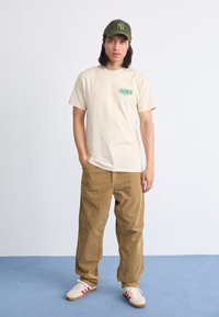 Camiseta de algodón beige con logo gráfico verde, combinada con pantalones de pana marrón. El modelo lleva una gorra verde y zapatillas blancas con acentos rojos.