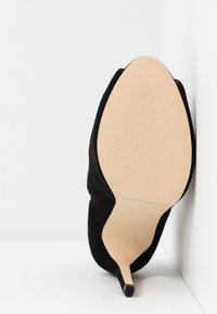 Musta samettinen korkeakorkoinen kengät, joissa on terävä kärki ja hoikka stiletto-korko. Vaalean värinen teksturoitu pohja, jossa on logo kohokuvioituna lähellä kantapäätä.